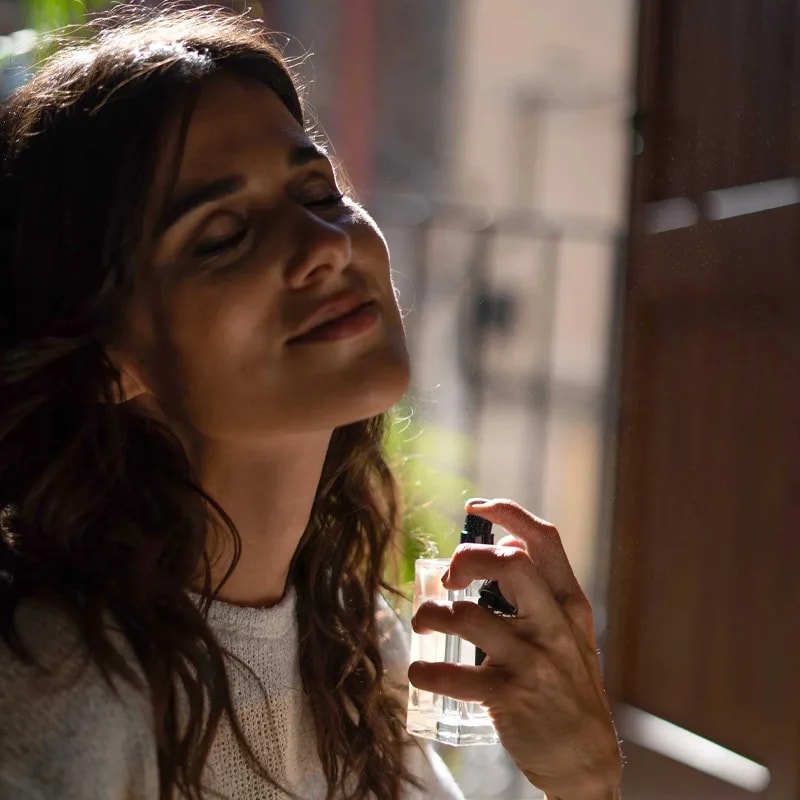 Woman in warm window light misting the Amber and Sandalwood fragrance towards her neck and hair
