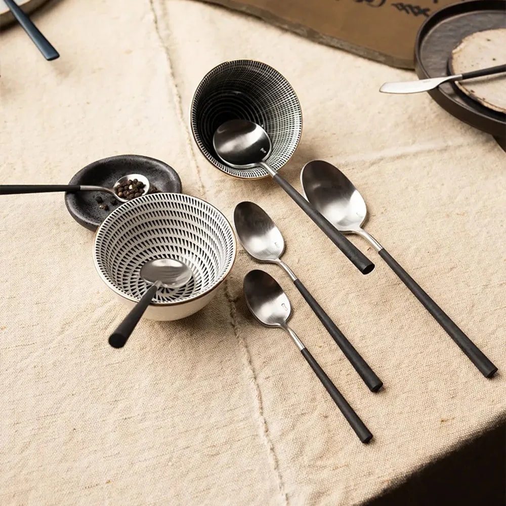 Angled table view of Neo Black spoons with matt black handles resting in striped bowls on linen