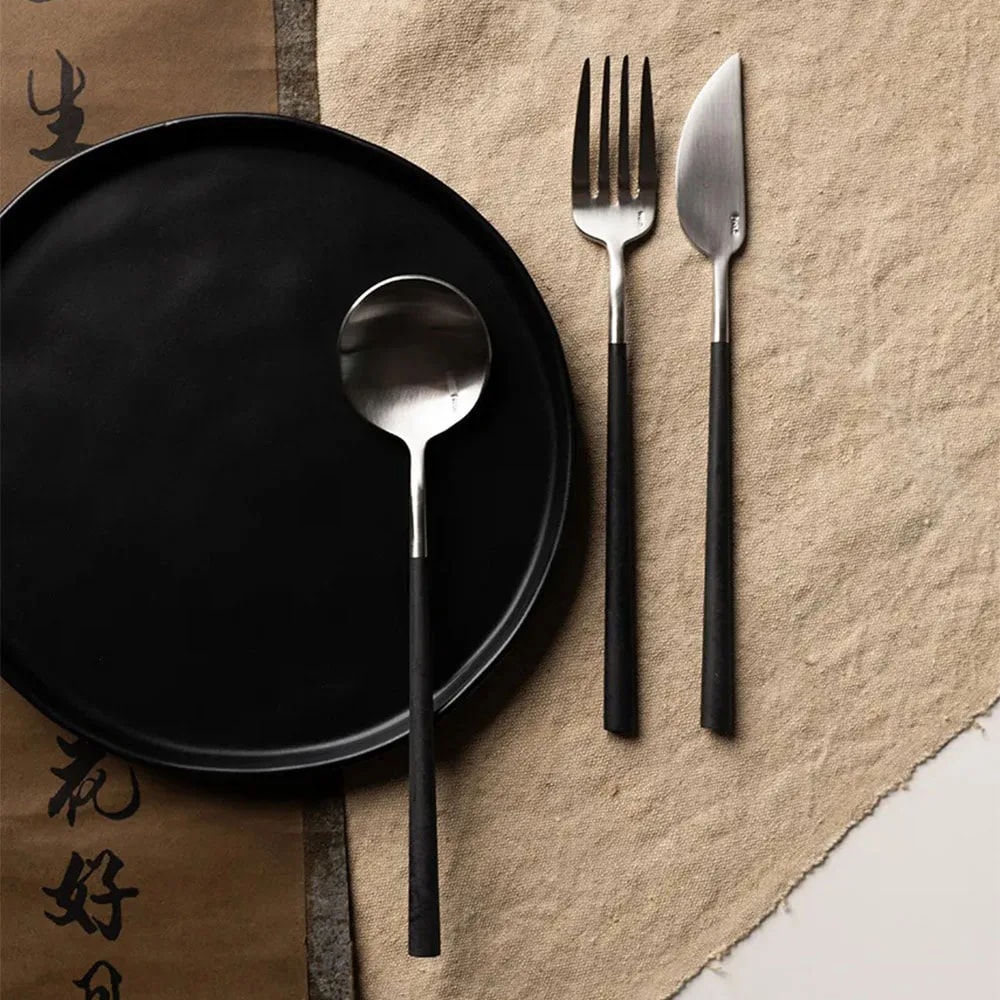 Place setting with a Neo Black spoon on a matt black plate next to fork and knife on a jute mat