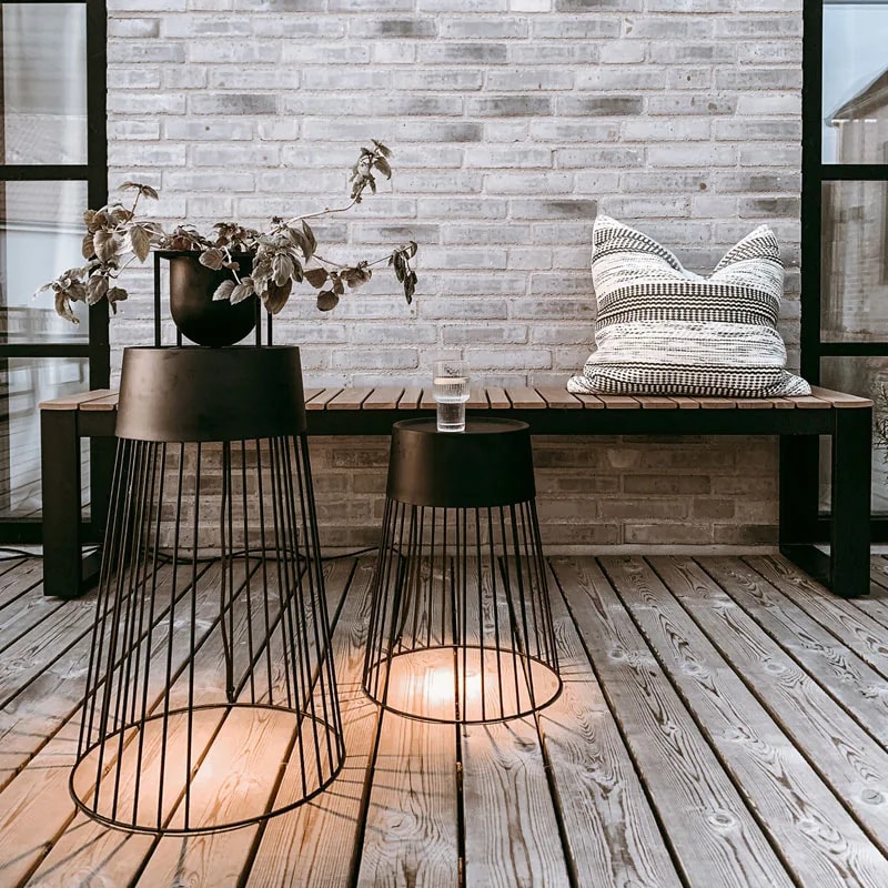 Two lit Koster cage floor lamps used as side tables on a decked terrace by a brick wall and bench