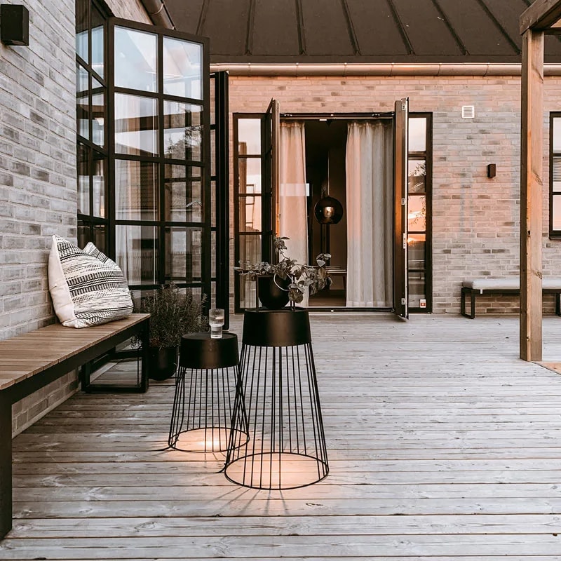 Koster cage floor lamps glowing on a wooden terrace at dusk beside open doors and a bench