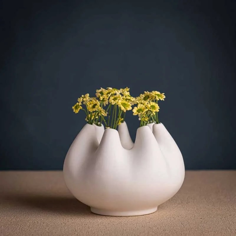 Rhizom 7s piccolo white vase holding yellow daisies on a sand-coloured surface, front view